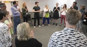 A group of adults in a workshop, standing in a circle with their palms pressed together in front of their chests.