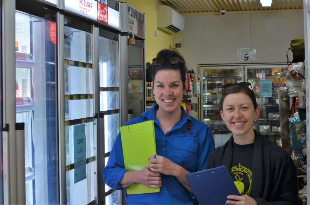 Rhiannon Hutchinson and Sara Bamford smiling as they carry clipboards in a store, Mimili
