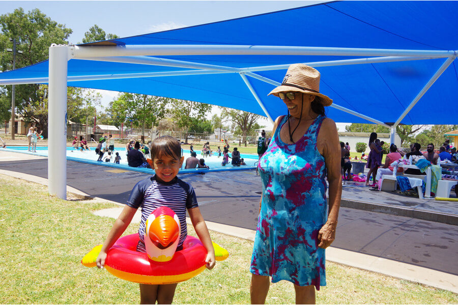 A child and adult enjoying themselves at Walgett Pool. Aly wears a red and yellow inflatable ring in the shape of a bird around his waist.