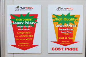 Two advertisements on the wall of Mai Wiru Regional Stores Council Aboriginal Corporation advertise “Lower prices, High quality” on a range of meat, fruit and vegetables. A legend at the bottom of the posters reads, “Healthy food, Healthy families.”