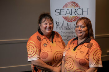 the Study of Environment on Aboriginal Resilience and Child Health (SEARCH) research officers Kym Slater and Sandra Banks, stand smiling next to a SEARCH banner.