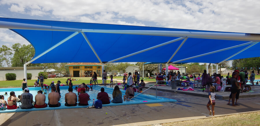 People enjoying Walgett Pool. Some are in the water while many others sit on the edge under a long blue shade stretching the length of the pool.