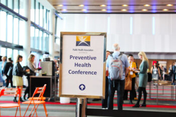 People network over coffee in a conference foyer. A sign with the logo of the Public Health Association of Australia (PHAA) reads "Preventive Health Conference".