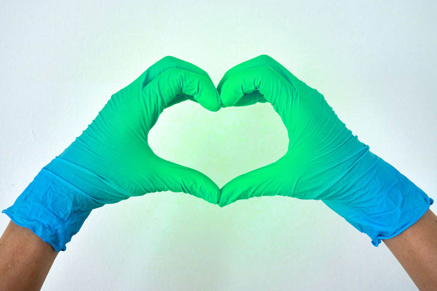 Closeup of hands in blue medical gloves forming a heart shape with the fingers. The heart is radiating green light.